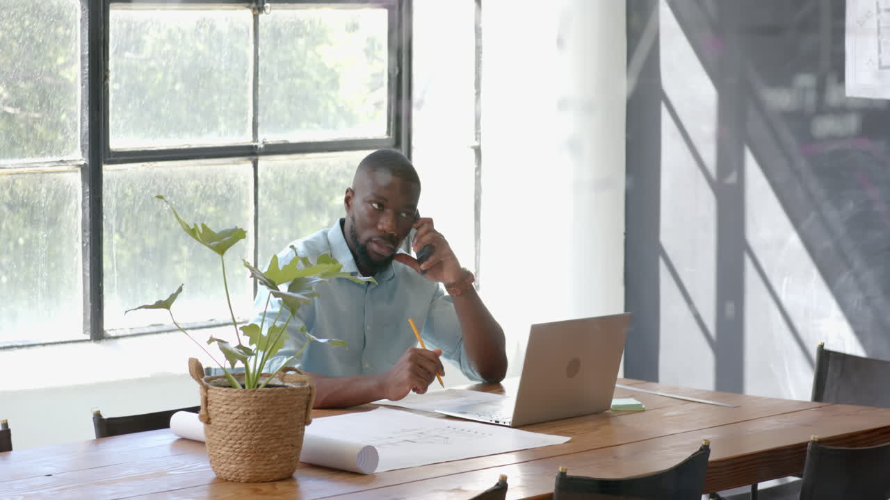 Talking on smartphone, man working on laptop and taking notes in office