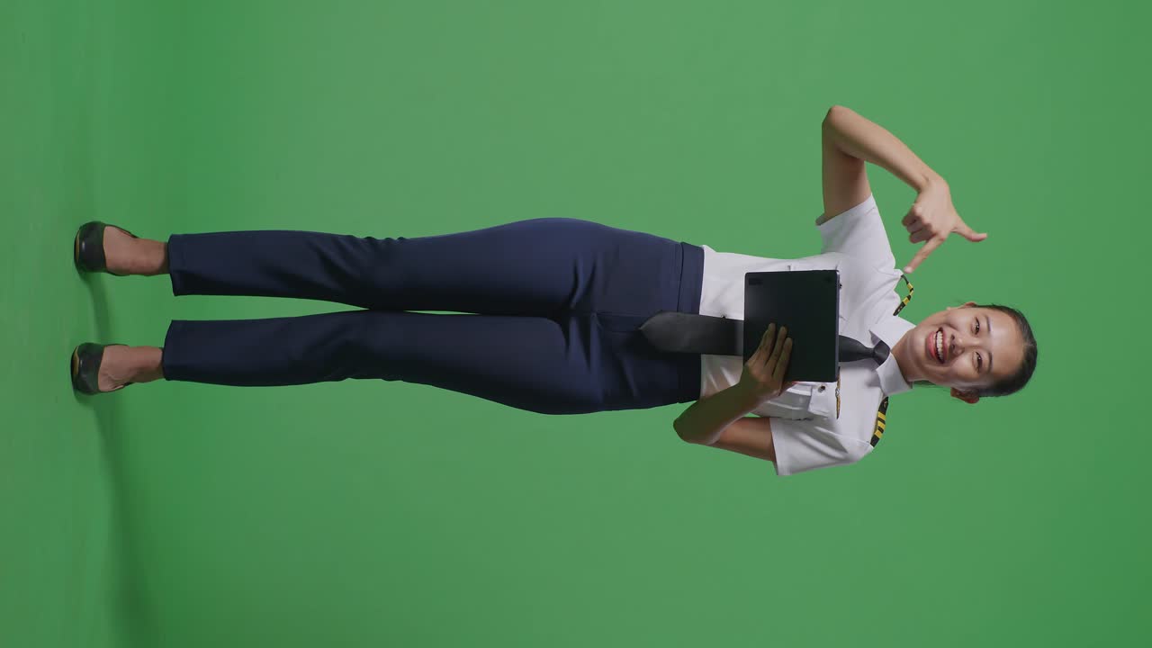 녹색 스크린 배경 스튜디오에 서 있는 동안 손에 태블릿을 가리키며 미소 짓는 아시아 여성 조종사의 몸이 가득 차 있습니다.