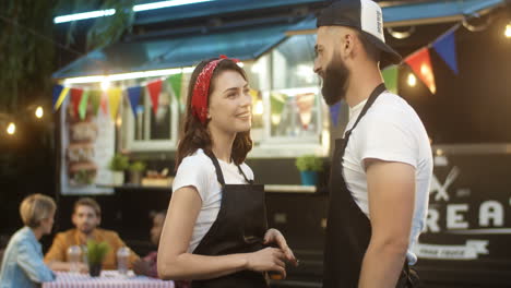 Couple-Of-Cheerful-Young-Couple-Of-Male-And-Female-Waiters-Talking-Outdoor-In-Festive-Food-Track-In-Park