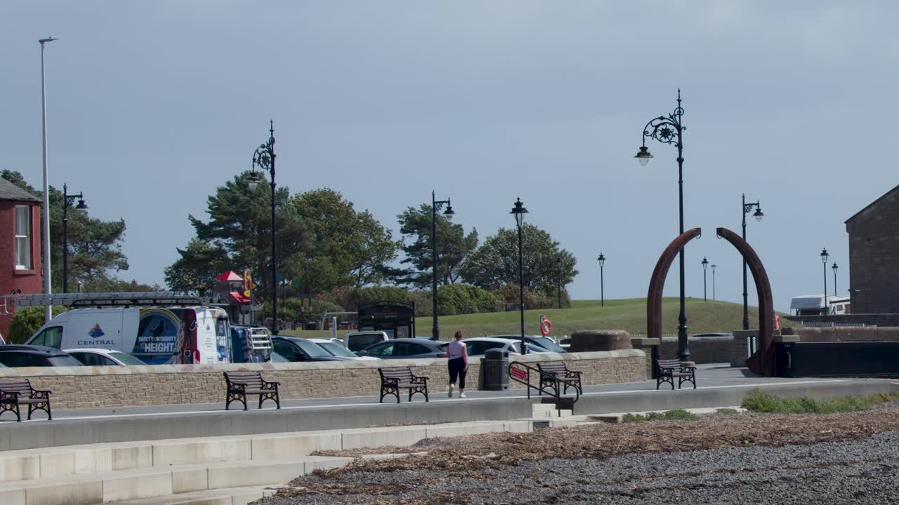Two people walk along a coastal promenade under overcast skies, distant buildings visible