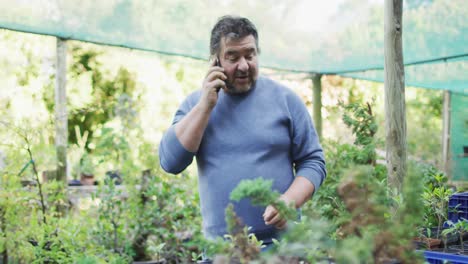 jardinero caucásico chequeando plantas y usando un teléfono inteligente en el centro de jardinería