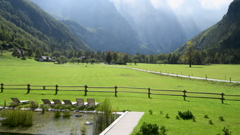 panoramic view in logarska valley, slovenia, green meadows with forest and high mountains in background, natural swimming pool with sunchairs in front