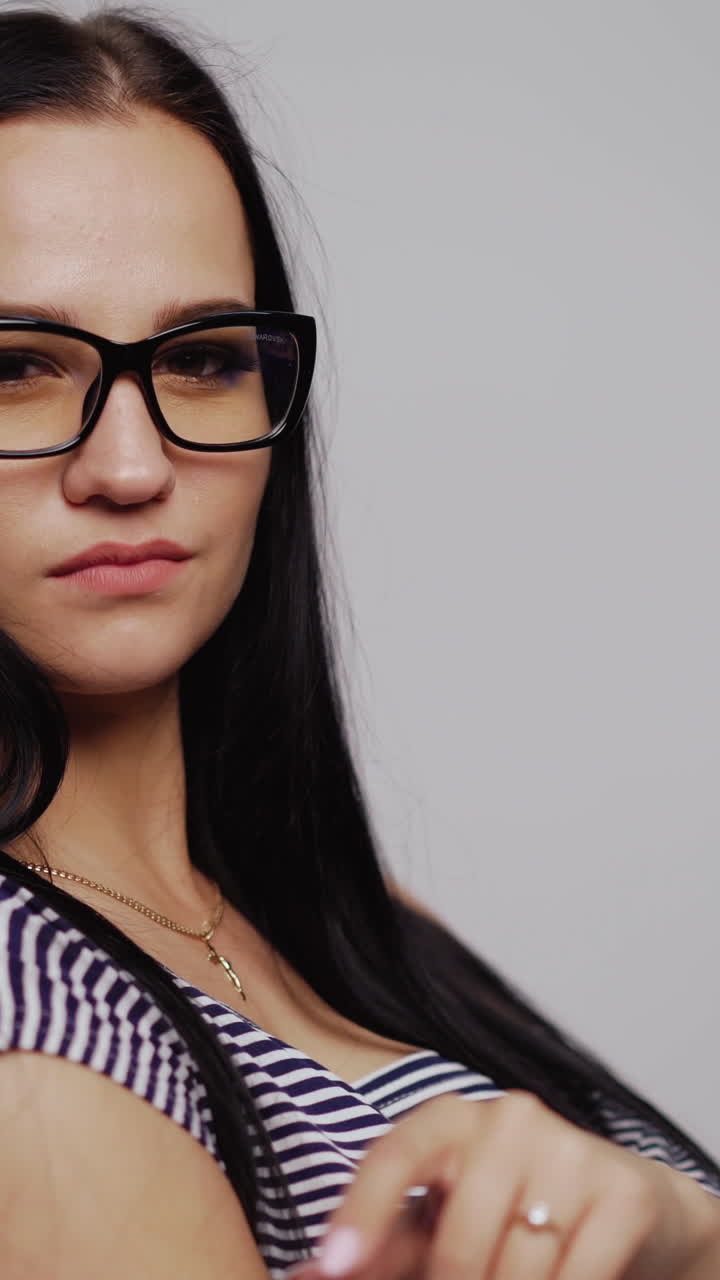 Serious young girl tries on fashionable eyeglasses in black rim over the light background. Attractive woman puts on new eyewear. Close-up Vertical video
