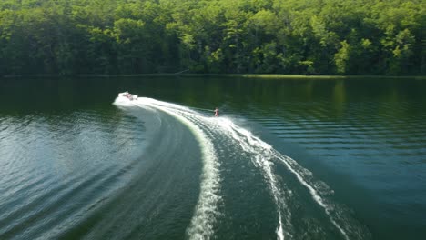 with numerous lush, green trees and few houses in the background, a woman in skis being carried by a speedboat at a blue lake is seen