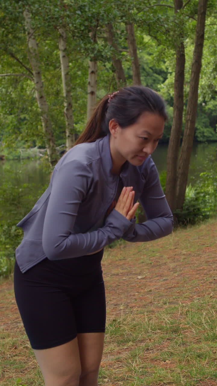 Vertikal video av kvinne i sportsklær som gjør yogastrekker på matte i skogen ved en innsjø eller elv, nyter naturens fred og skjønnhet, filmet i sanntid