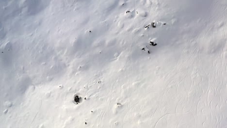 Drone-shot-looking-straight-down-and-dollying-backwards-onto-snow-field-with-rocks-and-ski-tracks,-in-La-Plagne-France