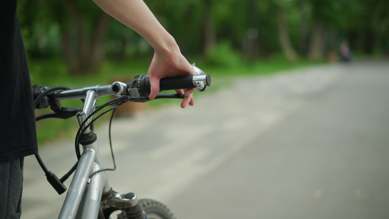 vista parcial de alguien con camiseta negra caminando de cerca con su bicicleta a lo largo de un camino tranquilo y pacífico, con el enfoque en su mano sosteniendo el manillar de la bicicleta, la carretera está rodeada de vegetación