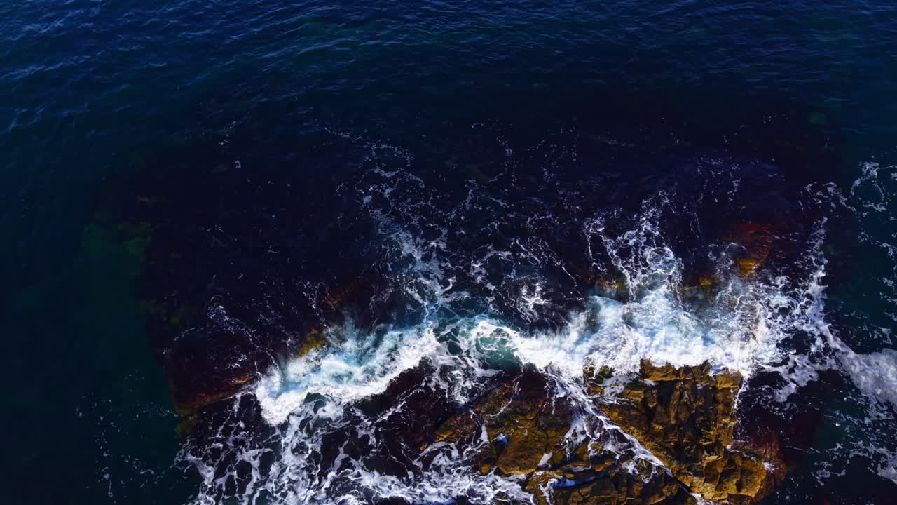 Waves crash dramatically over rugged rocks as the sun sets gently over the horizon, creating a stunning display of nature's beauty and power