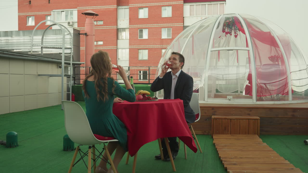 Cena en un iglú en la azotea con una pareja blanca, cúpula de burbuja íntima en la azotea, mantel rojo y centro de mesa floral, mujer con vestido verde sirviendo champán, hombre en traje levantando la copa, tintineo y risas