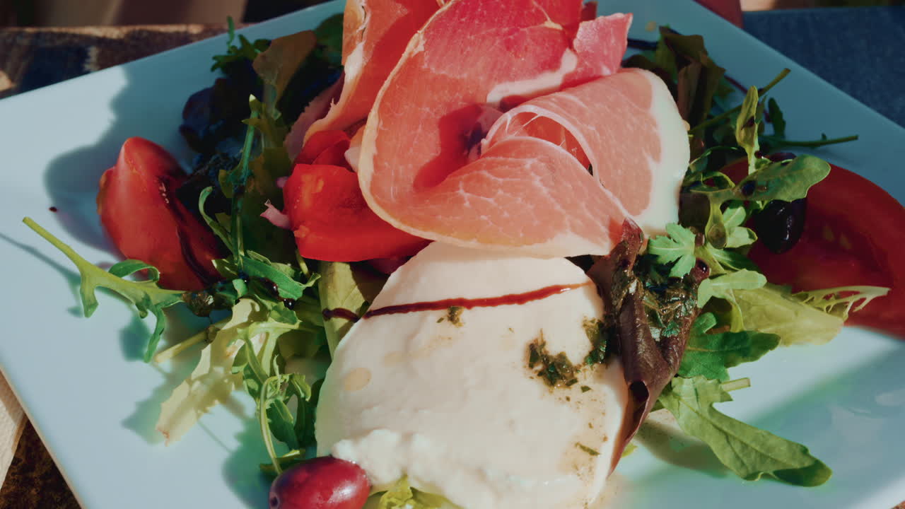 Close up of a gourmet salad with fresh mozzarella, cured meat slices, tomatoes, olives and mixed greens on a white plate in bright daylight
