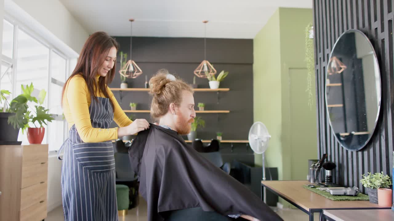 Happy caucasian female hairdresser putting cape on male client at hair salon, in slow motion