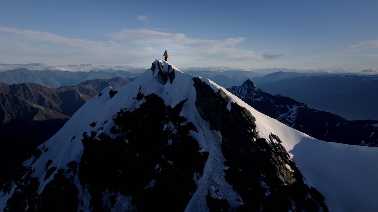 Un maestoso alpinista si erge in cima a una montagna innevata nelle selvagge distese della natura, con enormi montagne e valli gigantesche sullo sfondo