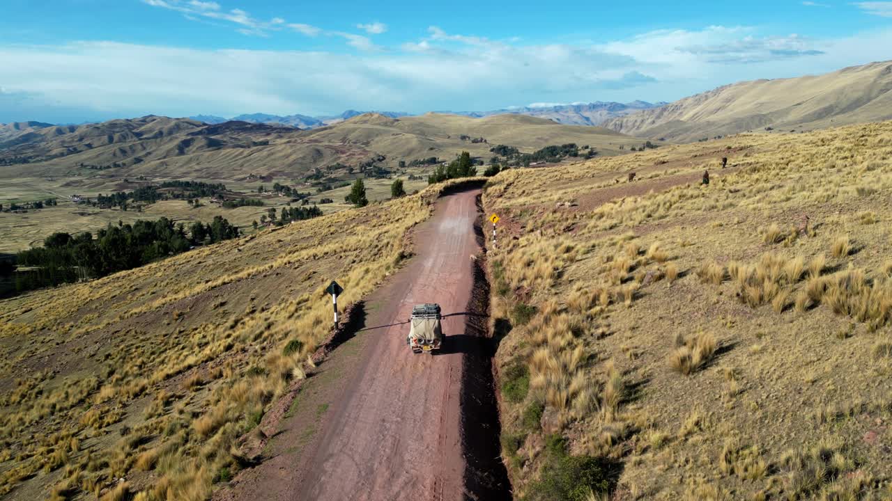 Aerial footage tracking a small vehicle driving through the high Andes. The road winds between golden hills and distant ridgelines