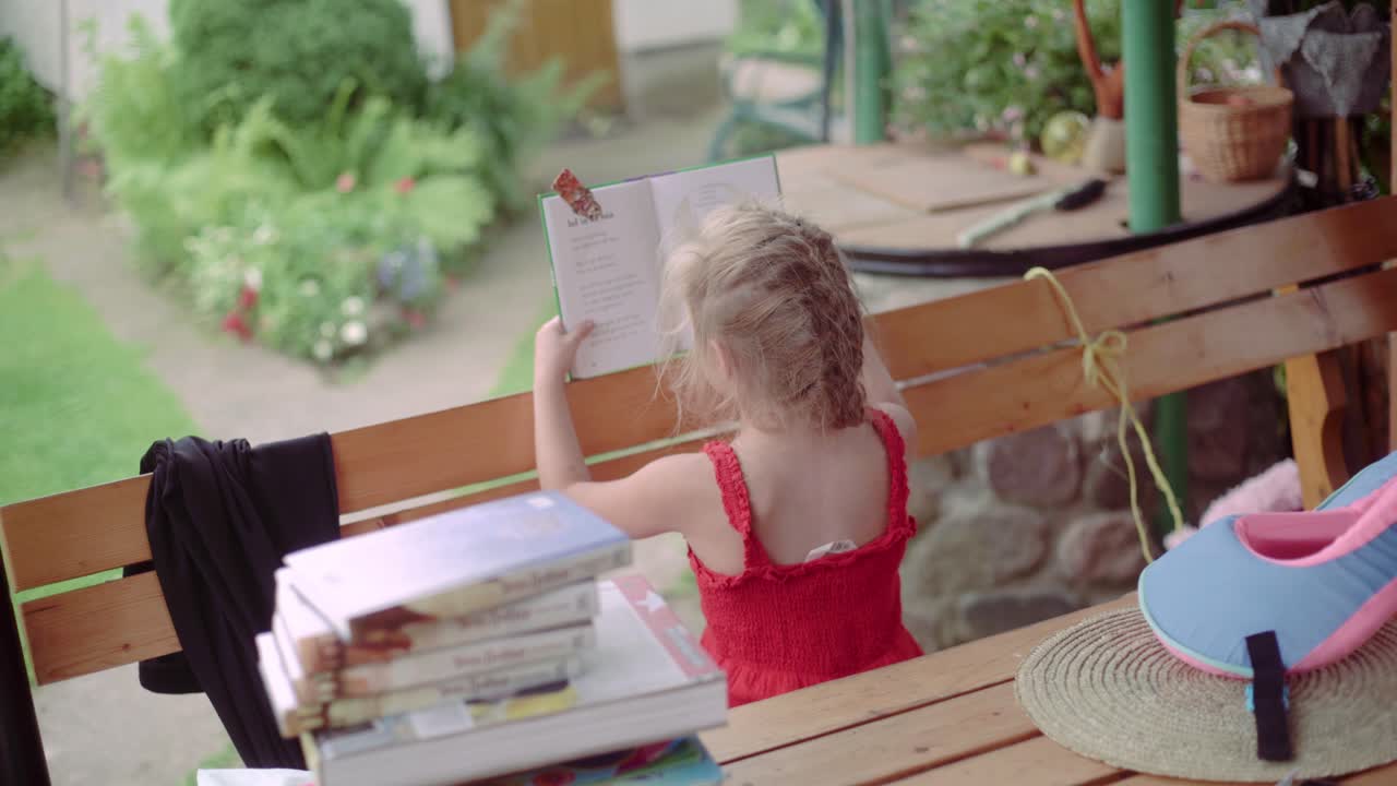 Cute little blonde girl reading children's books outdoors, View from the back