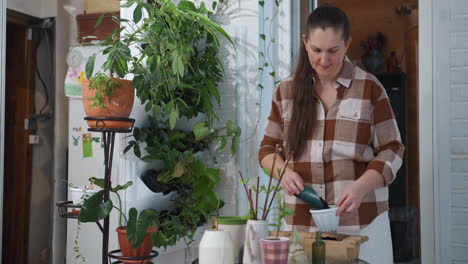 serene woman with focused face pours soil into white planter cup beside wooden container on indoor table surrounded by gardening tools and green plants in peaceful home gardening setting