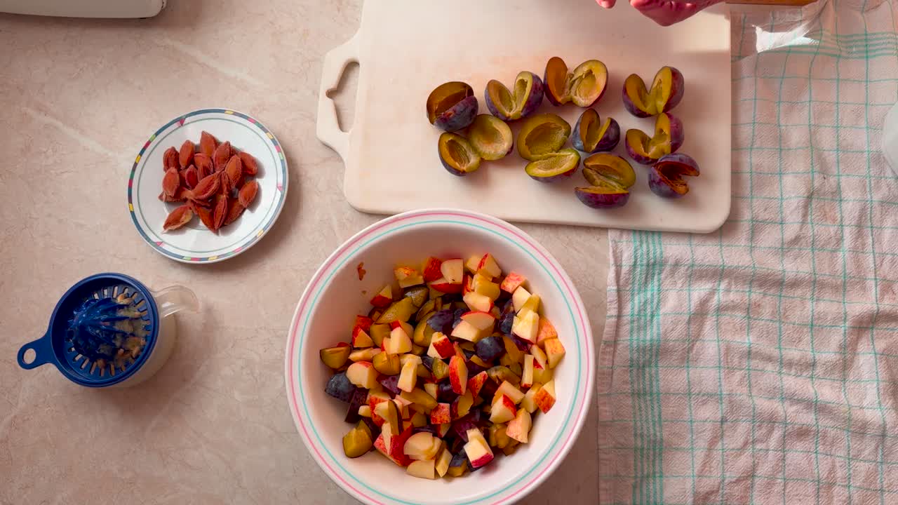 A person's hand is seen cutting plums, the pits are placed on a plate, and cut fruit is in a bowl, tilt down shot