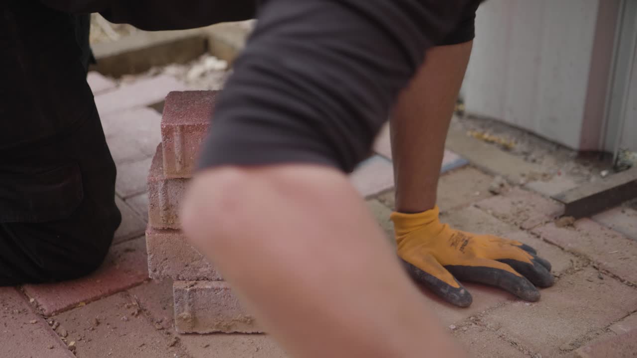 A construction worker in gloves carefully stacks bricks, picking them up from the ground. This shot captures the hard work and precision involved in construction and landscaping.