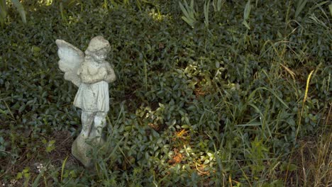 Small-stone-angel-statue-in-a-green-garden-on-a-bright-summer-afternoon-with-the-grass-blowing-gently-in-the-wind