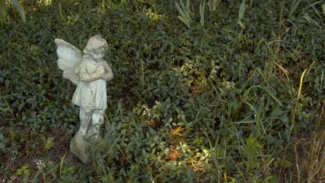 Kleine-Steinerne-Engelsstatue-In-Einem-Grünen-Garten-An-Einem-Hellen-Sommernachmittag,-Während-Das-Gras-Sanft-Im-Wind-Weht