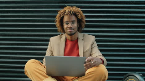 portrait of young african american man sitting on street with laptop