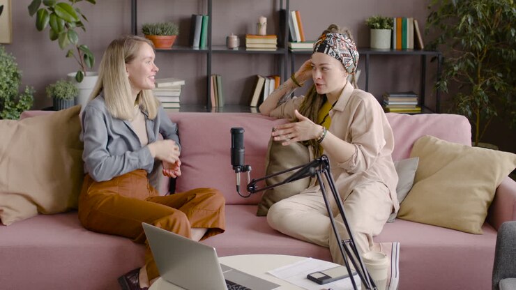 Two Women Recording A Podcast Talking Into A Microphone Sitting On Sofa In Front Of Table With Laptop And Documents 2