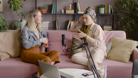 Two-Women-Recording-A-Podcast-Talking-Into-A-Microphone-Sitting-On-Sofa-In-Front-Of-Table-With-Laptop-And-Documents-2