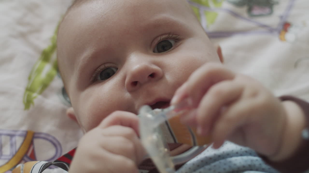 A four months old baby, lies on the coach with colorful bedsheet and playing with the baby's dummy toys, holding it in his hands
