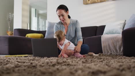 madre caucásica sosteniendo a su bebé usando una computadora portátil mientras trabaja desde casa