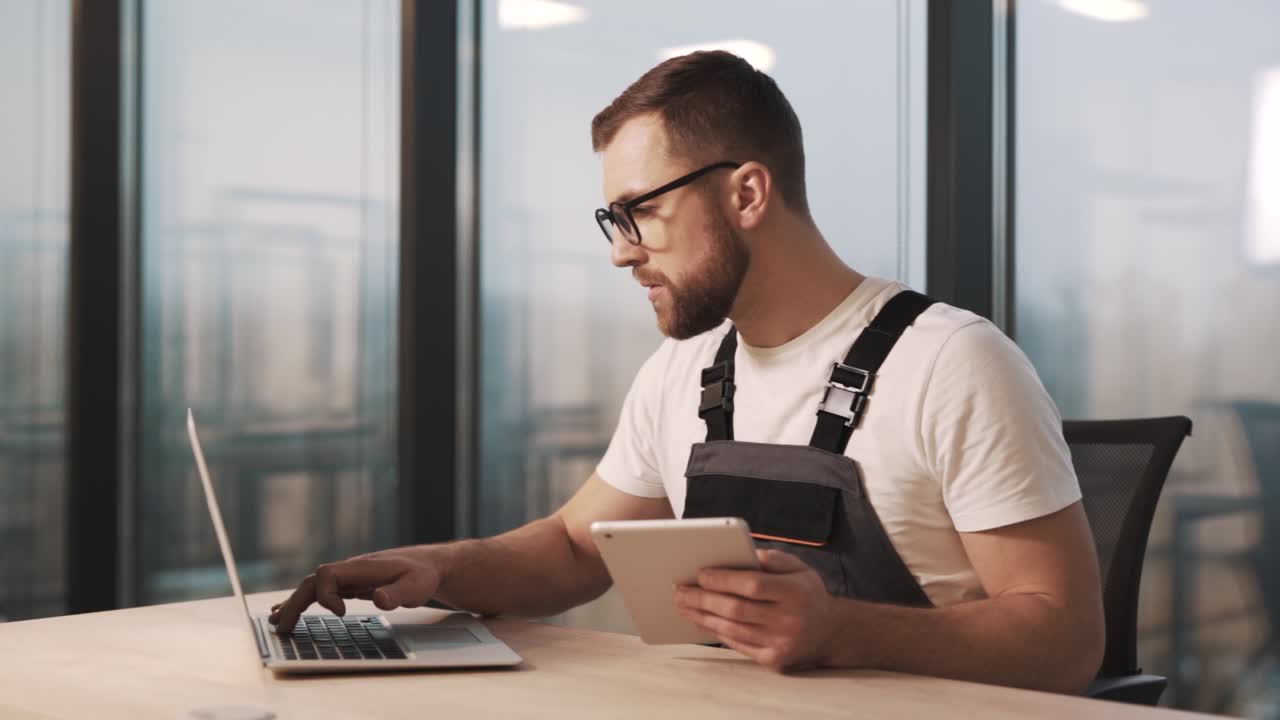 Portrait of technician in work uniform and glasses diagnoses laptop in modern office with large windows, repairs and installs new software from tablet