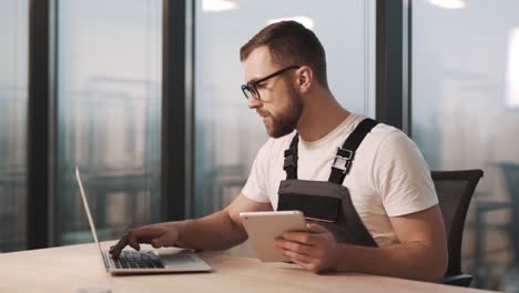 Portrait-of-technician-in-work-uniform-and-glasses-diagnoses-laptop-in-modern-office-with-large-windows,-repairs-and-installs-new-software-from-tablet