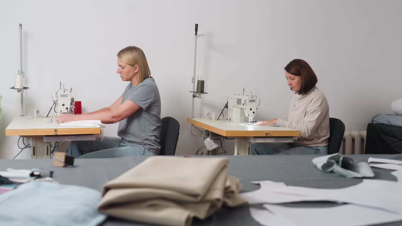 Stylists sewing white material at table with multiple fabrics in bright workshop, focusing on garment creation, showcasing skill, posture, synergy, and thorough craftsmanship for advanced finishing