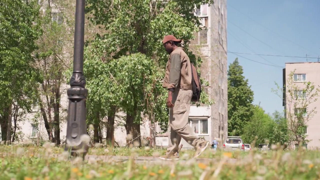 Man walks along grassy path in urban park wearing casual outdoor clothing and carrying backpack. scene features green trees tall apartment buildings with weathered facades parked cars
