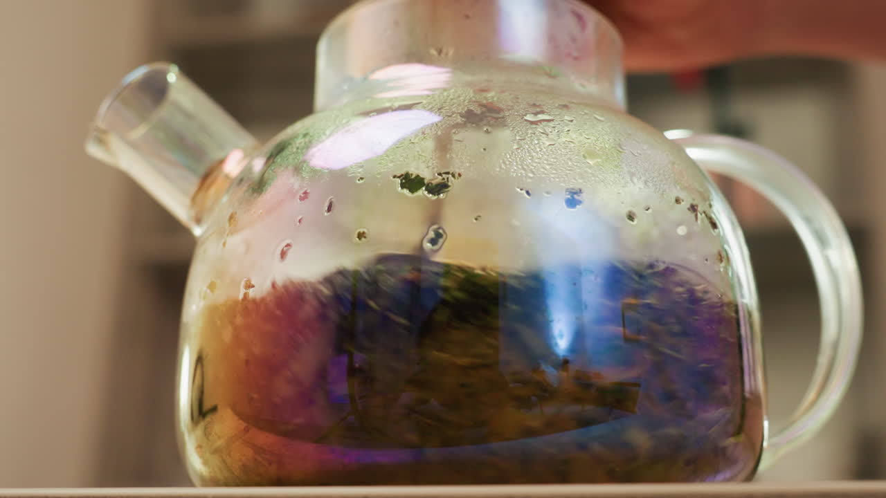 Teiera in vetro con infuso di erbe colorato, erborista casalinga che prepara una bevanda rilassante su un bancone di legno, condensa che si forma sul vetro, foglie di tè che turbinano nella calda luce mattutina, leggero vapore che sale