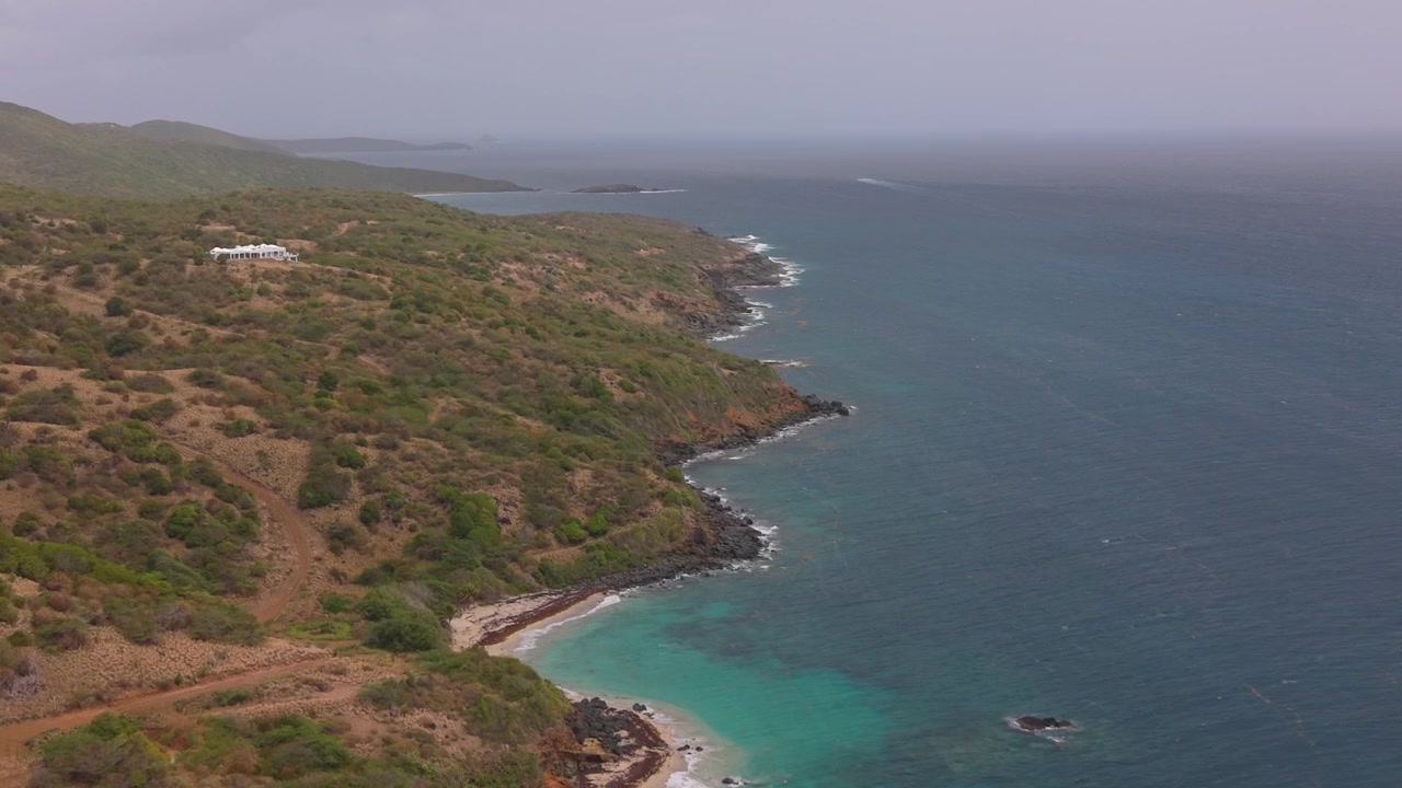 Una vista aerea che sorvola una costa tropicale, caratterizzata da colline verdi e scogliere rosse che incontrano acque oceaniche turchesi e limpide sotto un cielo velato