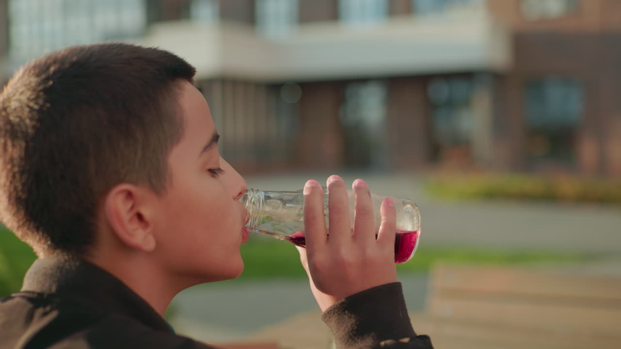 Side view of kid drinking fruit juice from glass bottle while holding sandwich outdoors at wooden table in warm sunlight, enjoying refreshing drink in relaxed urban atmosphere