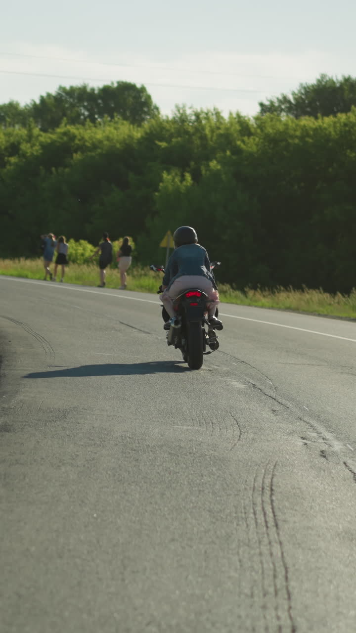 dos mujeres montando una bicicleta eléctrica alrededor de una curva, con una vista de personas caminando por el lado de la carretera y un coche que se acerca desde la dirección opuesta, la carretera está rodeada de vegetación