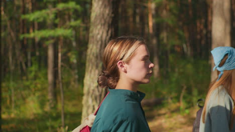 two women walking through sunlit forest, one with red backpack and cloth draped over it, other with blue headscarf and backpack, sunlight filters through trees, reflecting softly on their faces