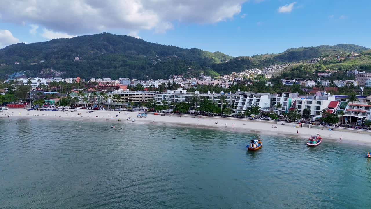 Drone footage captures vibrant Patong Beach with boats and scenic coastline under clear skies in Phuket, Thailand