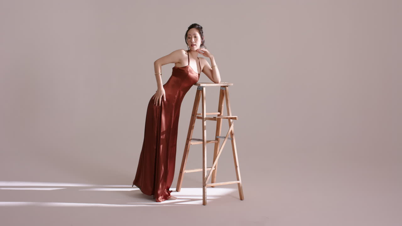 Photographer cueing Asian female model leaning on ladder in studio showing burgundy gown