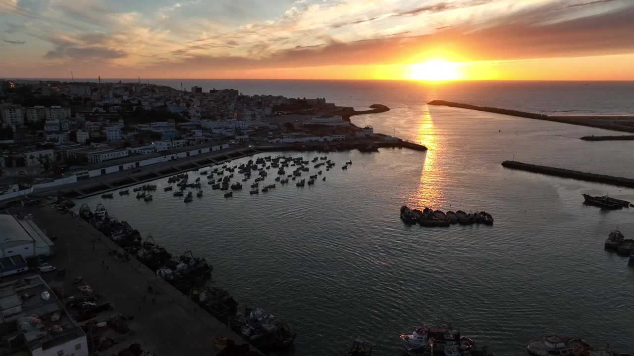 Gün batımında Asilah balıkçı limanının geniş havadan görünümü; yoğun küçük tekne filosu, liman dalgakıranları, kıyı şehri ve Atlantik suları üzerinde yansıyan altın ışık görülüyor.