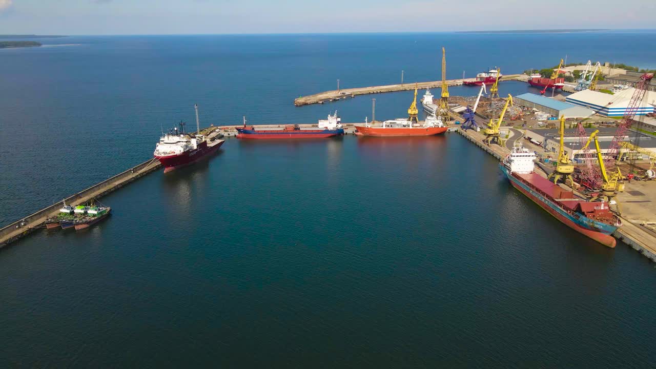 Aerial drone footage orbiting and flying over a dock harbor with large metal cargo and trading ships docked on the pier during a summer sunny day in Paljassaare with blue baltic sea ocean and horizon