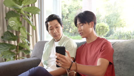 asian father and teenage son sitting on couch, son showing something on smartphone