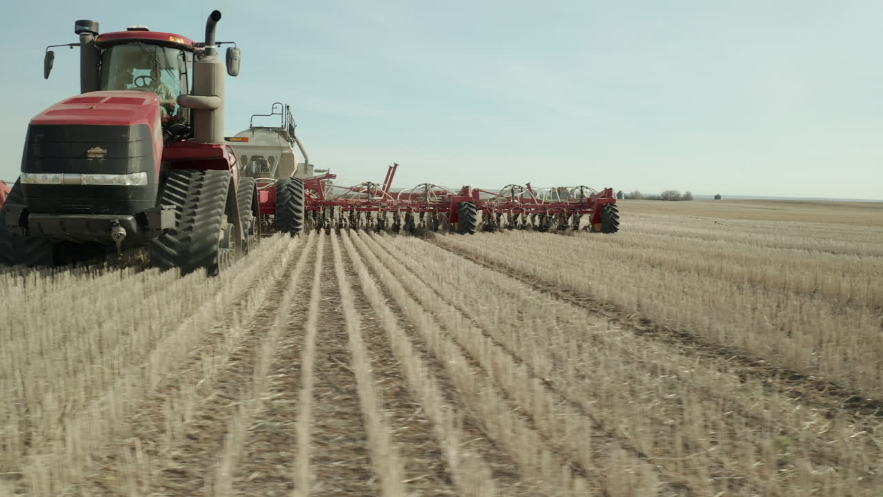 emocionante vuelo lateral bajo vista de cerca de la máquina tractora de siembra de granja roja que viaja en hileras planas de cultivos en un día soleado, vanguardia, saskatchewan, canadá, aéreo