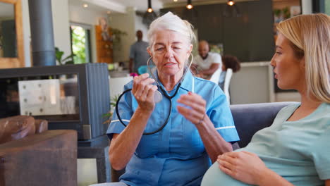 Senior-Midwife-With-Stethoscope-Visiting-Pregnant-Woman-At-Home-With-Family-In-Background