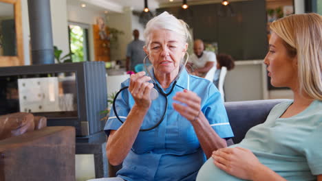 senior midwife with stethoscope visiting pregnant woman at home with family in background