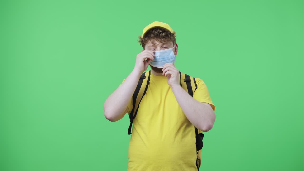 카메라를 바라보고 의료 마스크를 착용하는 젊은 남성의 초상화. 녹색 화면에 포즈를 취하는 테르모 가방과 함께 노란색 배달 택배 유니폼을 입은 은 머리 남성 59.97fps