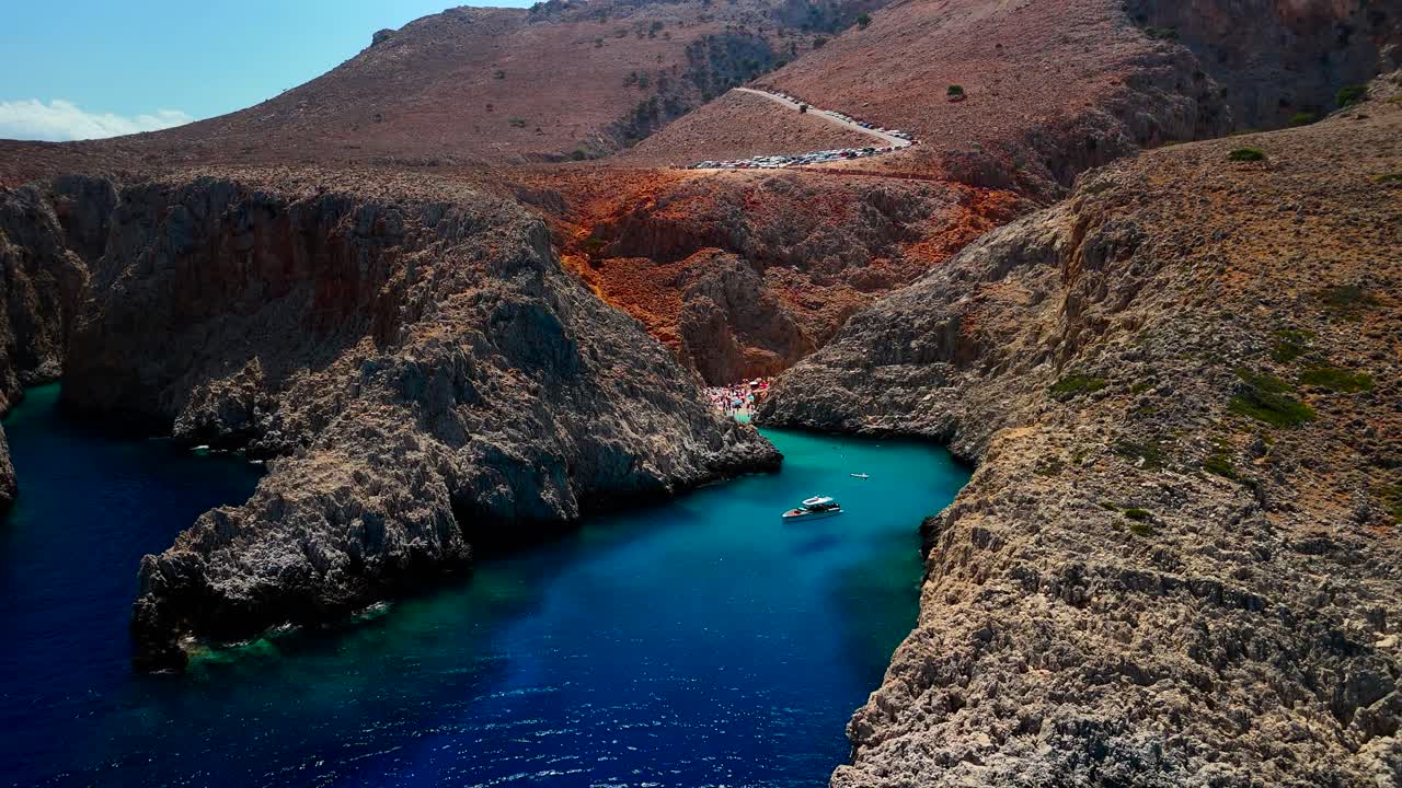 Visitors explore a stunning cove with bright blue waters nestled between steep rocky cliffs. The warm summer sun illuminates the area's natural beauty, making it perfect for relaxation