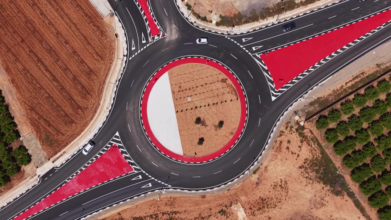 Top-down view of a large road roundabout with bold red-painted entry zones, white circle center, surrounding dry farmland and sparse trees with two cars visible