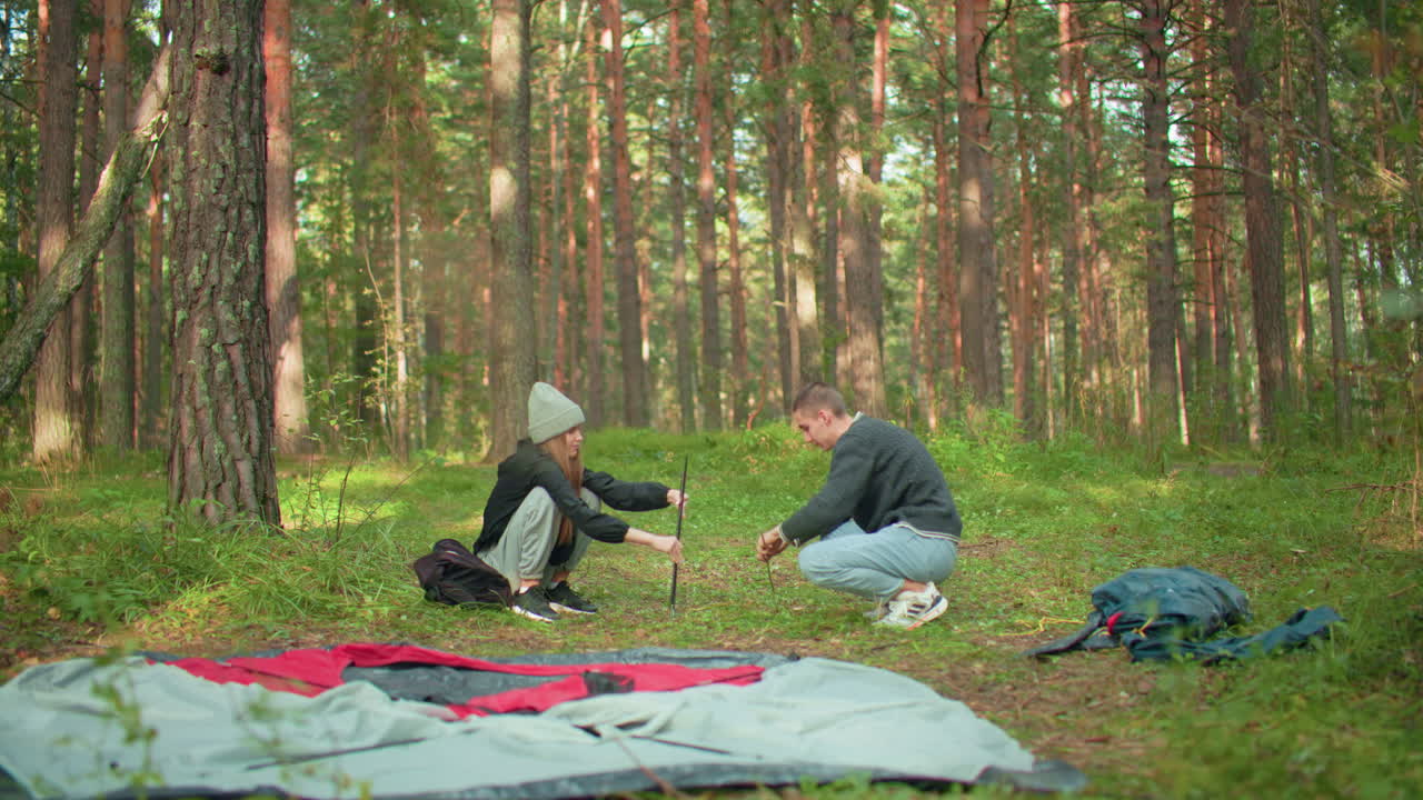 Lovers squatting together preparing tent for forest camping as lady hands over pole to her partner surrounded by scattered camping gear, dry leaves and backpacks