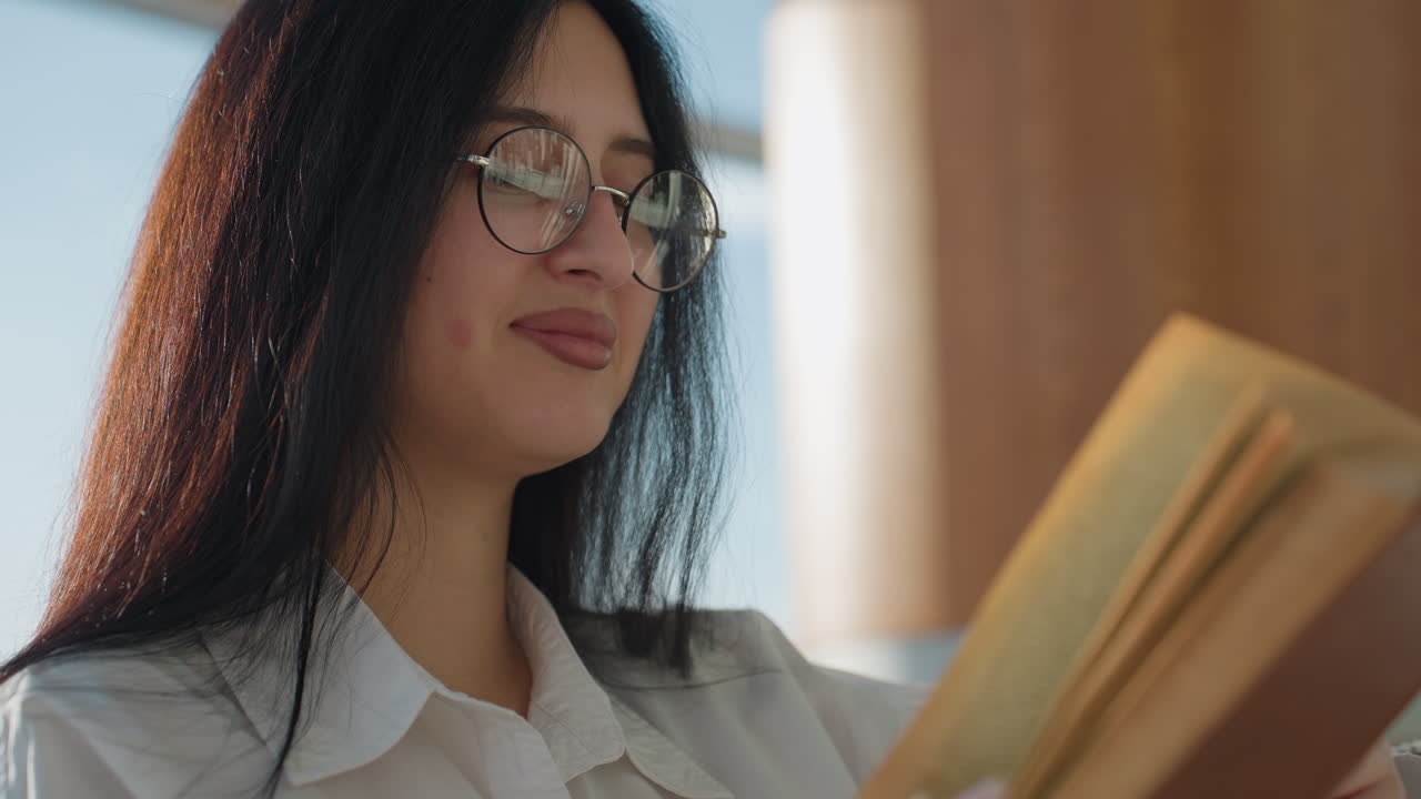 Elegant woman with glasses smiles subtly while reading novel in bright, modern setting with urban skyline in background, capturing serene intellectual moment in soft daylight near tall glass windows